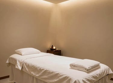 A wide shot of a luxurious South Asian / Indian spa room with beige walls, a professionally prepared massage bed with high-quality white linens, and soft ambient gold lighting creating a peaceful mood.