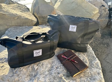 Two black Night Supply Co. duffel bags on rocks beside leather journal and pen.
