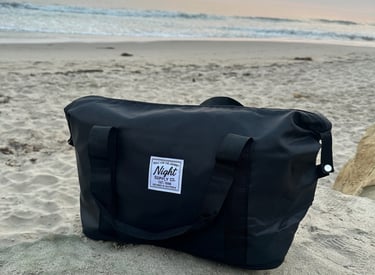 Black Night Supply Co. duffel bag on beach sand with sunset sky in background