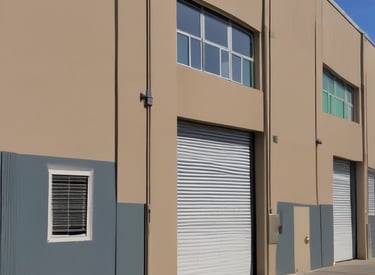 A person is painting on an easel outdoors on a tiled, wave-patterned sidewalk. Nearby, there are potted plants, chairs, and an iron gate attached to a building. The painting depicts a waterfront scene.