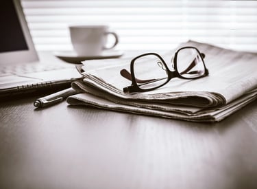 A desk with a laptop, a pen, a cup of tea, and a newspaper with a pair of reading glasses on it.