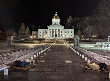 A nighttime picture of the lit Vermont capitol building. Two people are sleeping on the concrete.