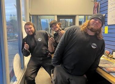 Three auto repair mechanics in black hoodies posing with thumbs up in a local tire shop.