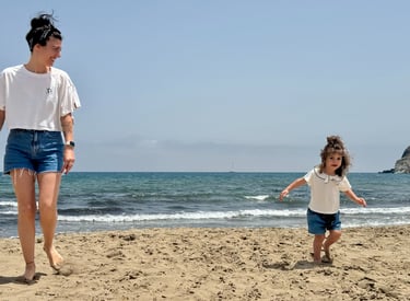 A mother and toddler daughter playing on a sandy beach by the ocean on a sunny day.