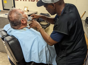 A student barber using electric trimmers to groom a customer's beard in a barber college.