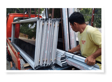 Two men unload glass display cases from a truck under a tree-lined street, preparing them for installation or transport.