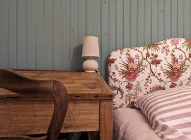 un lit avec une literie rayée, une tête de lit fleuri et un bureau d'écolier avec une lampe