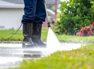 Person in rubber boots using a pressure washer to clean a wet outdoor concrete surface near grass and flowers