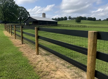 Black wooden horse fence with wire mesh on a green pasture farm with a barn and silo.