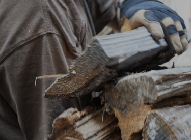 un homme en tee shirt gris porte pleins de bûches empilées dans ses mains.