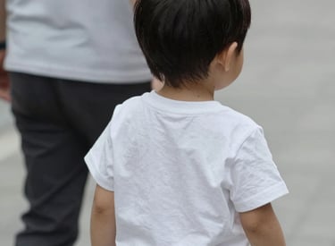 A person sits on a green yoga mat, wearing bright pink leggings and white socks. They hold two small jars, one with a green label reading 'CBD Balm' and the other lid removed, showing its contents. The person wears rings and has black-painted nails.