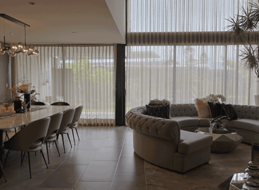 Sala-comedor con cortinas traslúcidas de doble altura, sofá en escuadra y mesa de mármol | KARÄNA