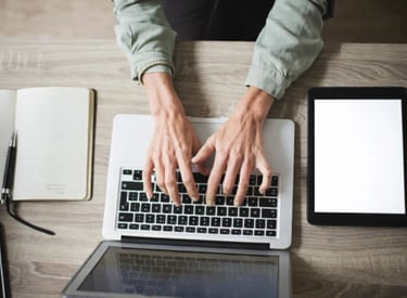 A copywriter typing on a laptop computer with a digital tablet and notebook on a desk.