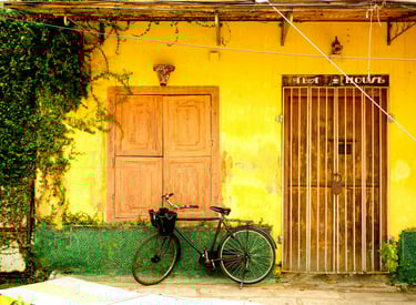 Bicycle parked in front of an old tea house with yellow walls and wooden doors, surrounded by lush green vines, Fort Kochi.