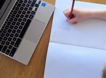 An 11+ student's hand writing math in a grid notebook next to a silver laptop learning at home