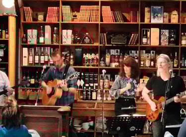 Live band performing acoustic music in a cozy bar with shelves of liquor bottles and books.