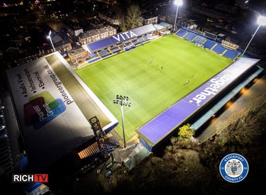 Aerial night view of Stockport County FC Edgeley Park stadium illuminated by bright floodlights.