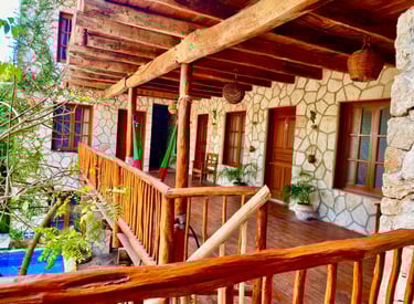 Rustic stone villa balcony with wooden beams, hanging hammocks, and tropical plants overlooking a pool.