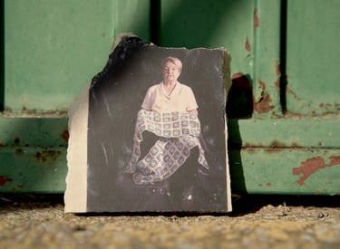 A vintage portrait of an elderly woman holding a patterned quilt on a stone fragment against a green metal door.