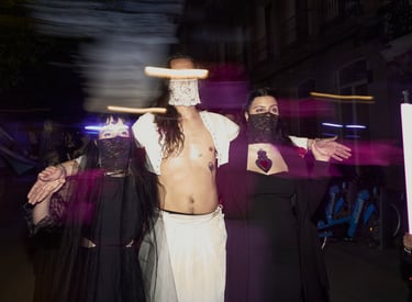 Performers in gothic avant-garde costumes and lace face veils pose during a nighttime city street event.