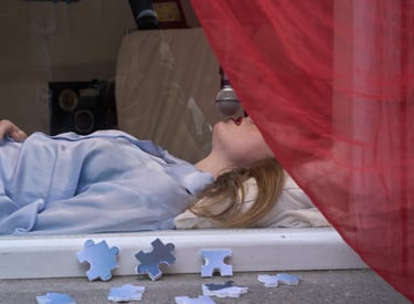 A woman lying behind glass with a microphone near her mouth, surrounded by red fabric and puzzle pieces.
