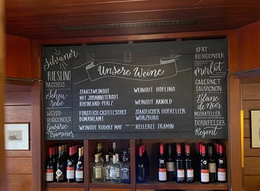 Handwritten wine menu on a chalkboard featuring German Riesling and Silvaner above shelves of wine bottles.