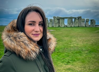 A woman in a green parka smiles in front of the prehistoric Stonehenge monument under a cloudy sky.