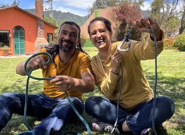 A smiling couple sits on a grassy lawn practicing rock climbing knots with a blue rope and carabiner.