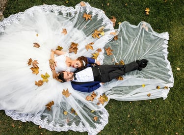 A bride and groom lying on a green lawn covered in autumn leaves and the white lace train of a wedding dress.