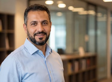 A smiling bearded businessman in a light blue button-down shirt standing in a modern office.