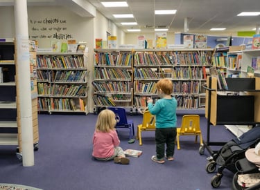 Toddlers exploring books in the children’s area of Saffron Walden Library, low shelves, colourful chairs & space for buggies.