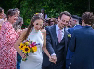 Smiling bride in a halter dress holding sunflowers walks with her groom at an outdoor wedding.