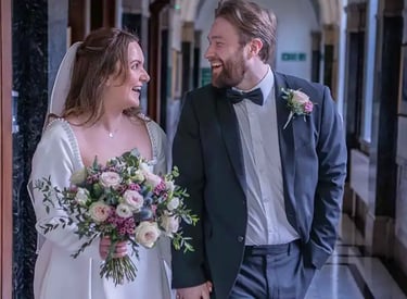 Smiling bride in white gown and groom in tuxedo walking down hallway with a floral bouquet.