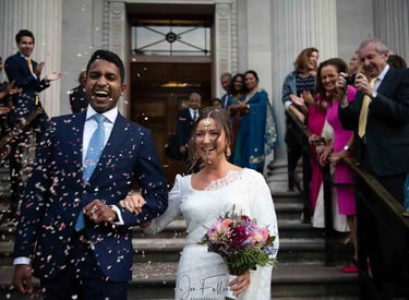 Smiling bride and groom walk through falling confetti after their wedding ceremony at a historic building.