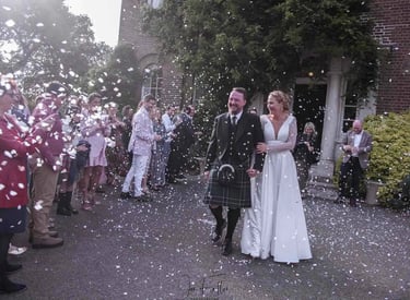 A bride and groom in a traditional kilt walk through falling confetti at an outdoor wedding.