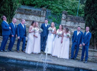 A happy wedding party in blue suits and white bridesmaid dresses posing by a garden fountain.
