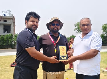 A man in a white polo shirt presents a sports award trophy to a coach on a sunny grass field.