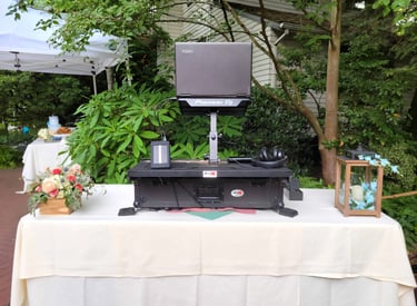 A DJ setup in an outdoor space decorated with flowers and white tablecloth to compliment the setting