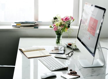 A bright workspace with keyboard, monitor, mouse, desk accessories, and flowers in a vase next to a window.