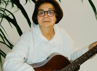 Diane Fischer playing an acoustic guitar indoors next to a green plant.