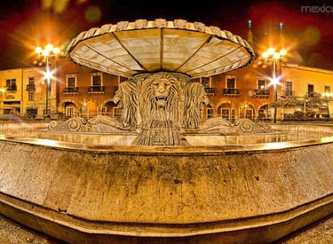 Illuminated lion fountain at night in a historic Mexican city plaza with glowing street lights.