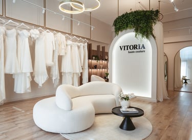 Luxury bridal boutique interior with elegant white wedding dresses, a curved bouclé sofa, and Vitoria Haute Couture branding.