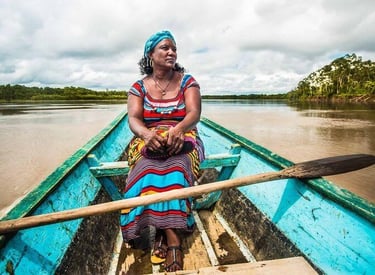 A woman in a colorful dress sitting in a wooden boat on a tropical river surrounded by rainforest.