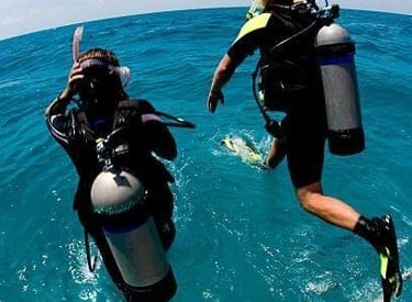 Two scuba divers jumping off a boat into clear blue ocean water with full dive gear.
