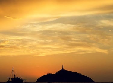 Silhouetted fishing boats on calm water at sunset with a lighthouse island on the horizon.