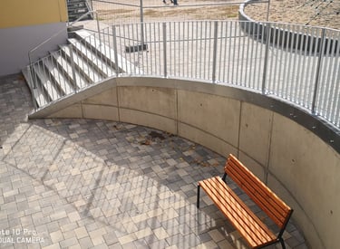 Modern outdoor courtyard with a wooden park bench, curved concrete wall, and metal railing stairs.