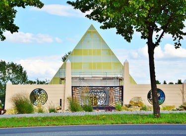Modern pyramid-shaped building with a sunburst gate and circular windows framed by lush green trees.