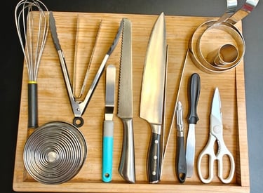 Professional chef tools neatly arranged on a bamboo board including a whisk, knives, tongs, pastry rings and kitchen scissors