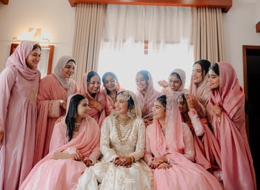 Smiling bride in white lehenga surrounded by bridesmaids in pink hijabs at a traditional Nikah ceremony.