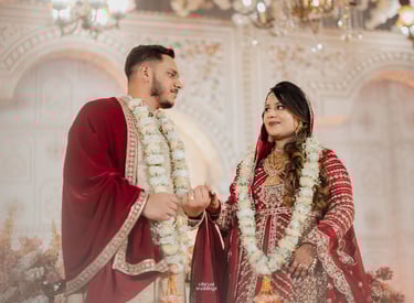 A couple in traditional red and white South Asian wedding attire posing under a chandelier.